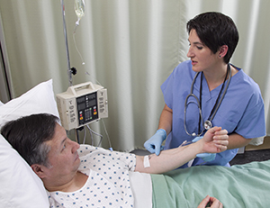 Hombre acostado en una cama de hospital con sonda intravenosa (i.v.) en el brazo. Una proveedora de atención médica revisa la sonda intravenosa (i.v.).